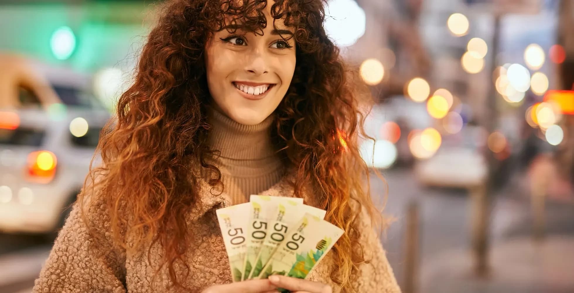 Junge Frau mit lockigen Haaren hält mehrere 50-Euro-Scheine in der Hand und lächelt, unscharfer Stadtverkehr im Hintergrund.